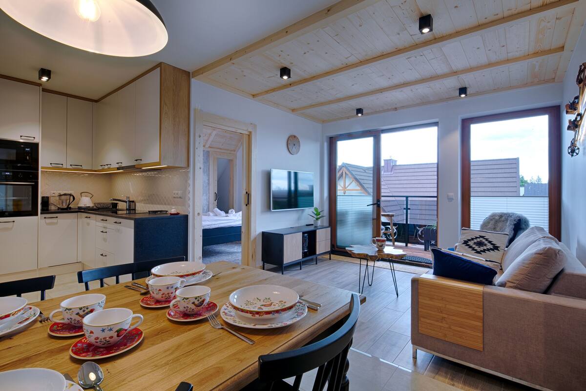 A modern kitchen with wooden cabinets, a dining table set with tea cups, and a living area with a sofa and a TV.
