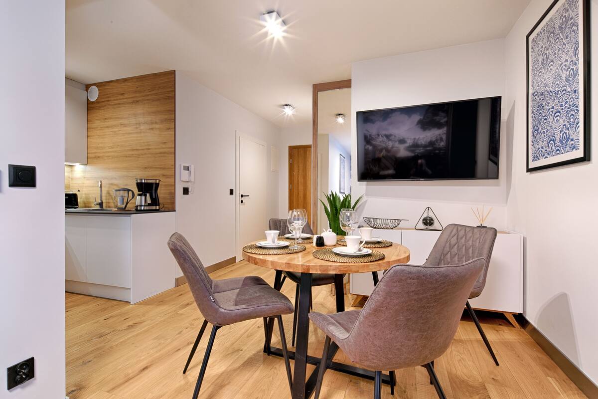 A modern dining area with a round wooden table, four grey upholstered chairs, and a large framed artwork on the wall.