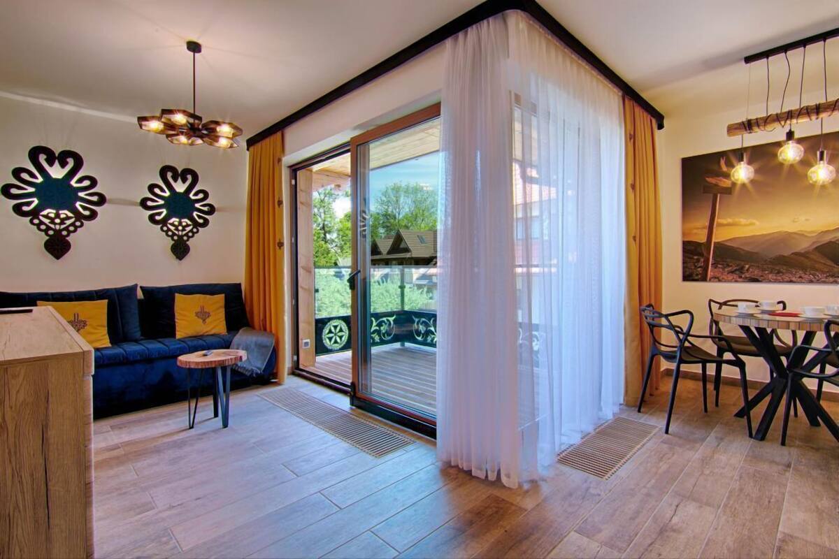 A modern living room with a sofa, a small table, and a dining area with chairs. There is a large sliding glass door leading to an outdoor balcony.