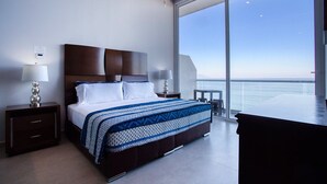 A modern hotel room with a large bed, two bedside tables, a nightstand, a desk, and a view of the sea.