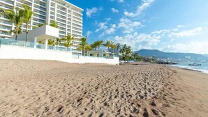 A beachfront hotel with a sandy beach, palm trees, and a clear blue sky.