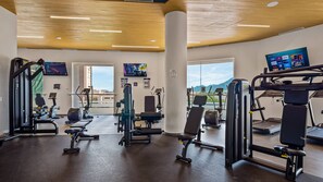 Et fitnesscenter med forskellig træningsudstyr, store vinduer og et fjernsyn, der viser træningsvideoer.