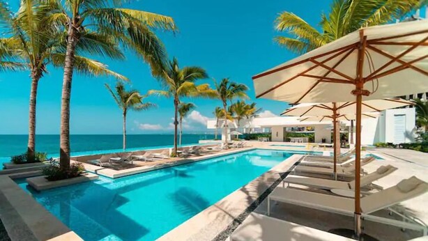 A pool area with lounge chairs and umbrellas, palm trees, and a view of the ocean.