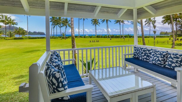 A covered outdoor seating area with white railings, blue patterned cushions, and a clear view of a well-maintained lawn and palm trees. - Hawaii Life Rental Presents Makana Nui (Hanalei)