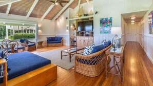 A spacious living area with a high ceiling, wooden beams, wicker furniture, a television, and a painting on the wall. - Hawaii Life Rental Presents Makana Nui (Hanalei)
