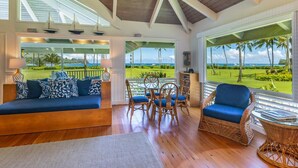 A spacious living area with a wooden ceiling, large windows offering a view of a golf course, a blue sofa, wicker chairs, and a small table. - Hawaii Life Rental Presents Makana Nui (Hanalei)