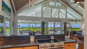 A kitchen with a large island, stainless steel appliances, and large windows offering a view of the outdoors. - Hawaii Life Rental Presents Makana Nui (Hanalei)