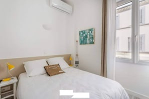 Un dormitorio con una cama, una mesita de noche, una lámpara, un cuadro y una ventana.