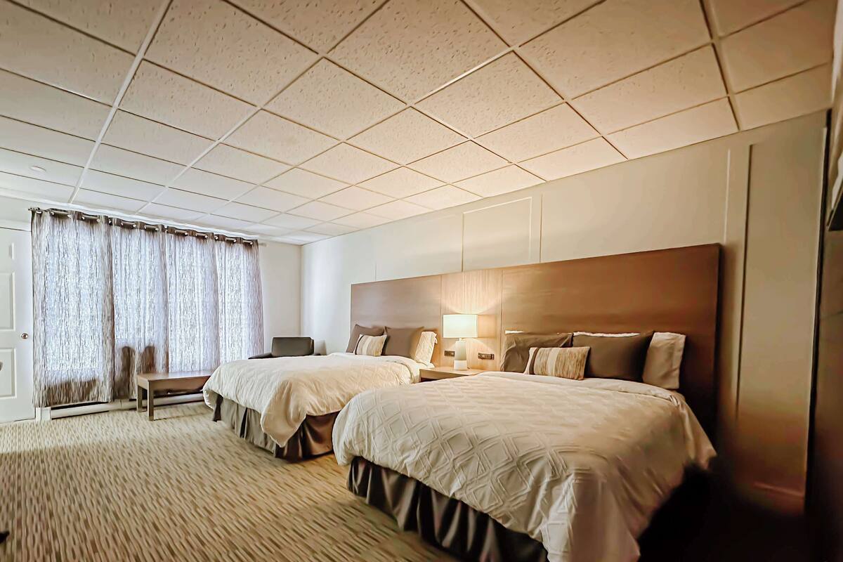 Quarto de hotel com duas camas, uma escrivaninha, uma cadeira e uma janela grande com cortinas.