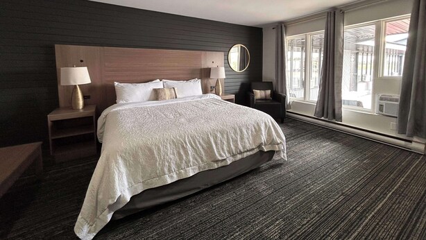 A hotel room with a large bed, two bedside tables with lamps, a round mirror, and a view of the outside through large windows.