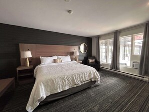 A hotel room with a large bed, two bedside tables with lamps, a round mirror, and a view of the outside through large windows.