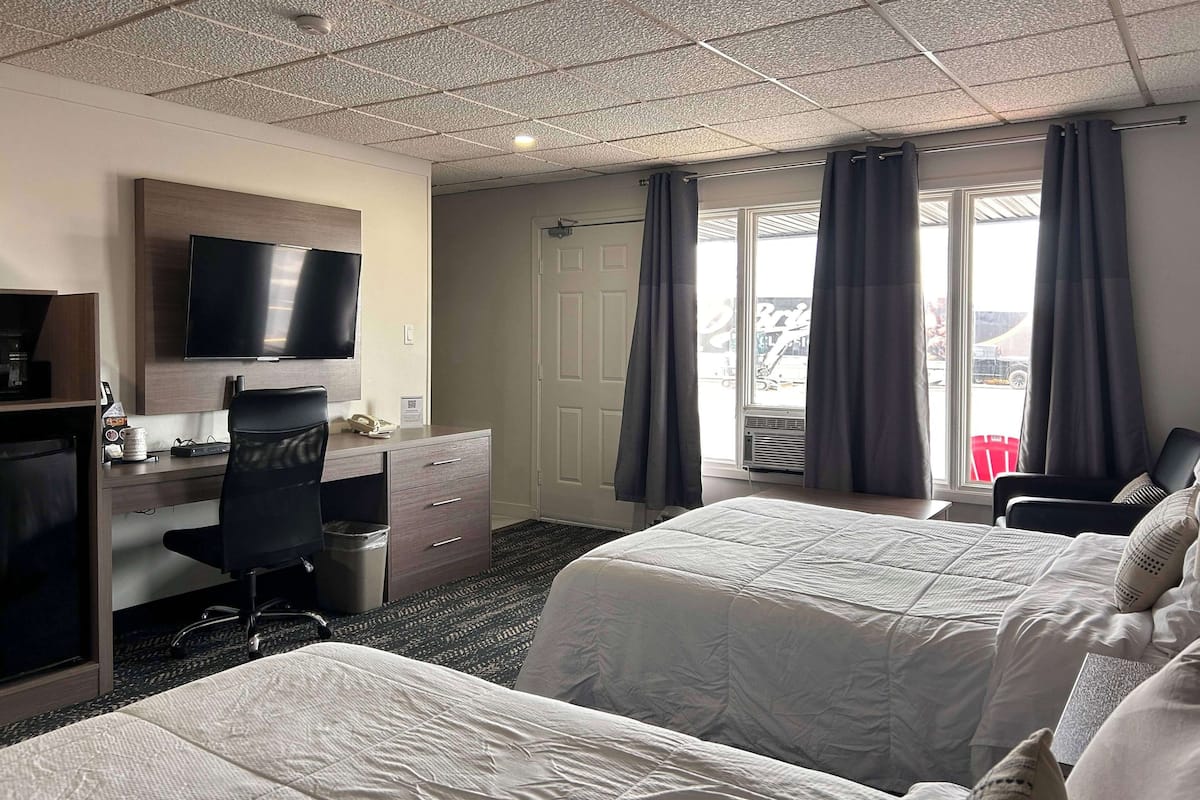 Quarto de hotel com duas camas, uma escrivaninha com cadeira, televisão e janela com cortinas.