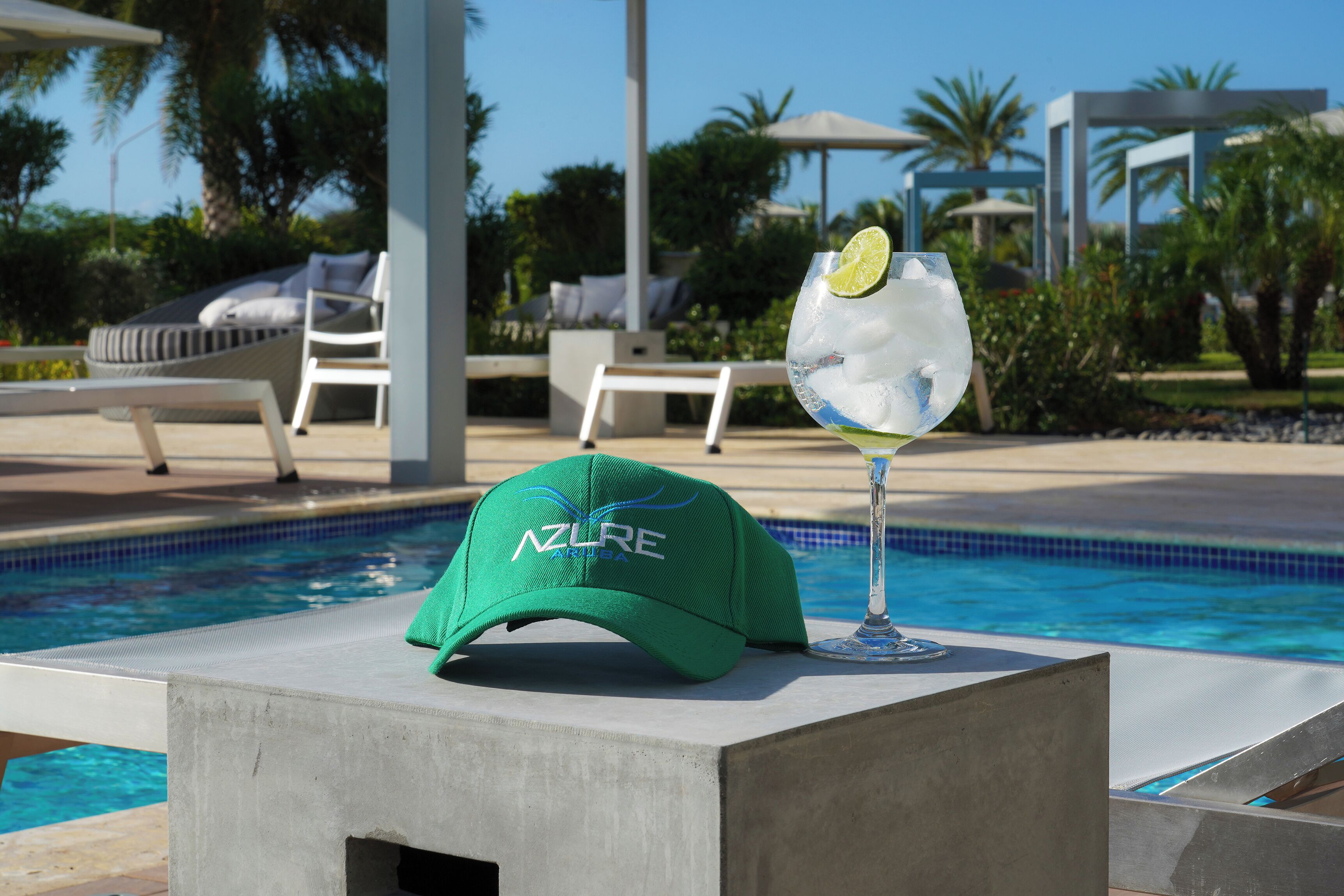 Zona de piscina con sillones reclinables, un bloque de concreto, un vaso con un trozo de lima y una gorra con el texto 'AZURE'.