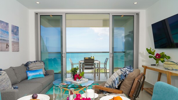 A modern living room with a balcony overlooking the sea, furnished with a sofa, dining table, and chairs. - Azure Beach Residences (Noord)