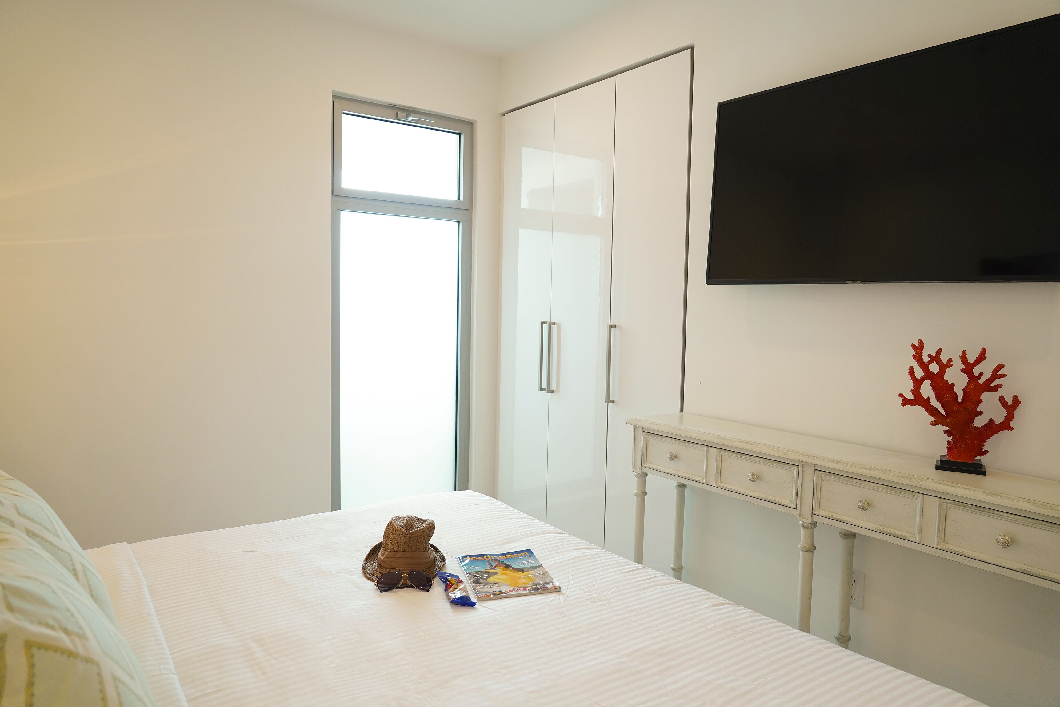 Un dormitorio con cama, televisor montado en la pared, armario y una pequeña mesita con cajones.