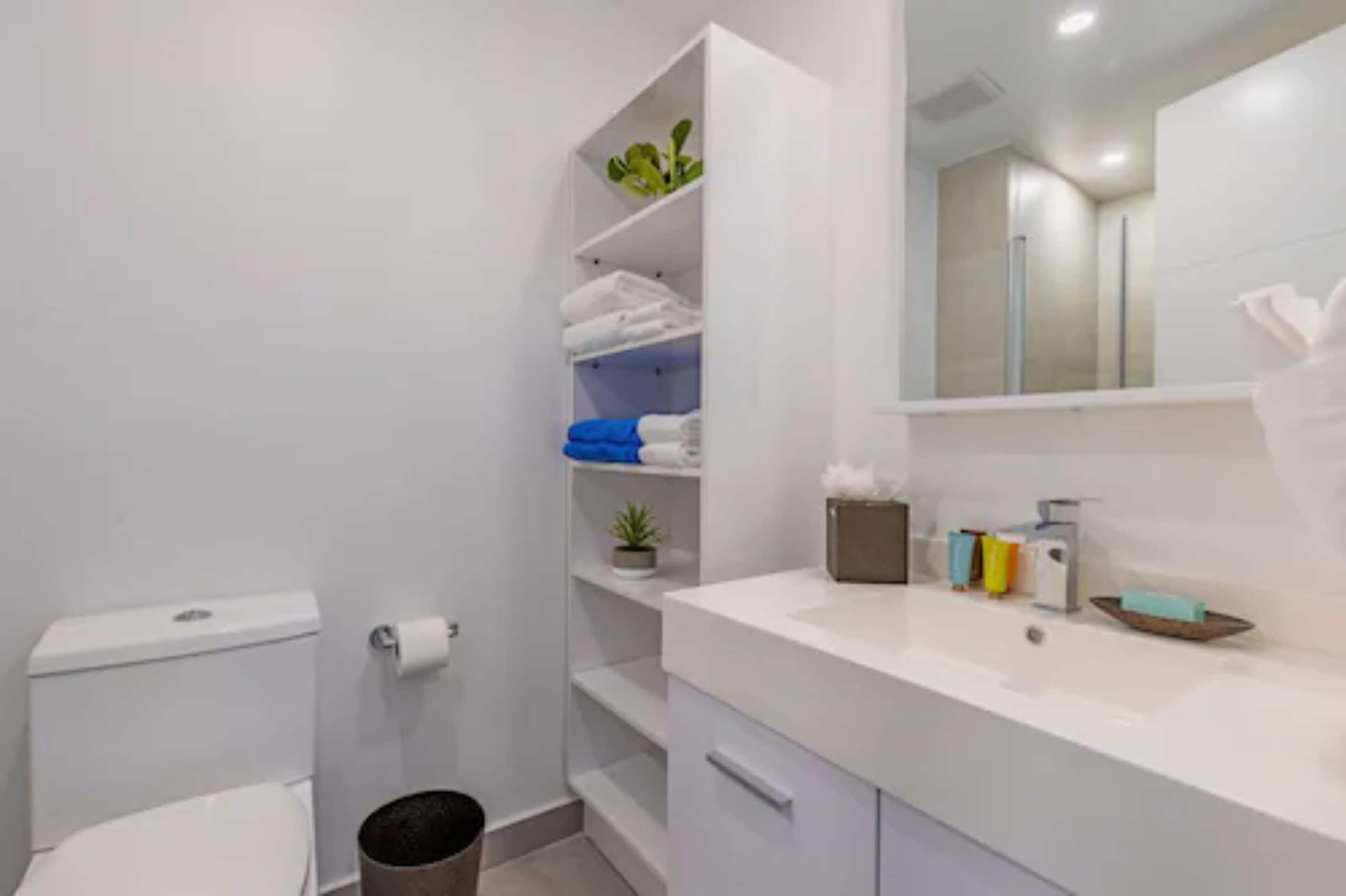 A modern bathroom with a toilet, a sink with a mirror, and a shelf with folded towels and a plant.
