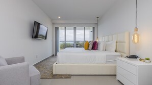 A modern bedroom with a large bed, a TV mounted on the wall, and a bedside table with a lamp. - Azure Beach Residences (Noord)