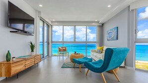 A modern living room with a large TV, a sofa, a blue armchair, and a view of the ocean through large windows. - Azure Beach Residences (Noord)