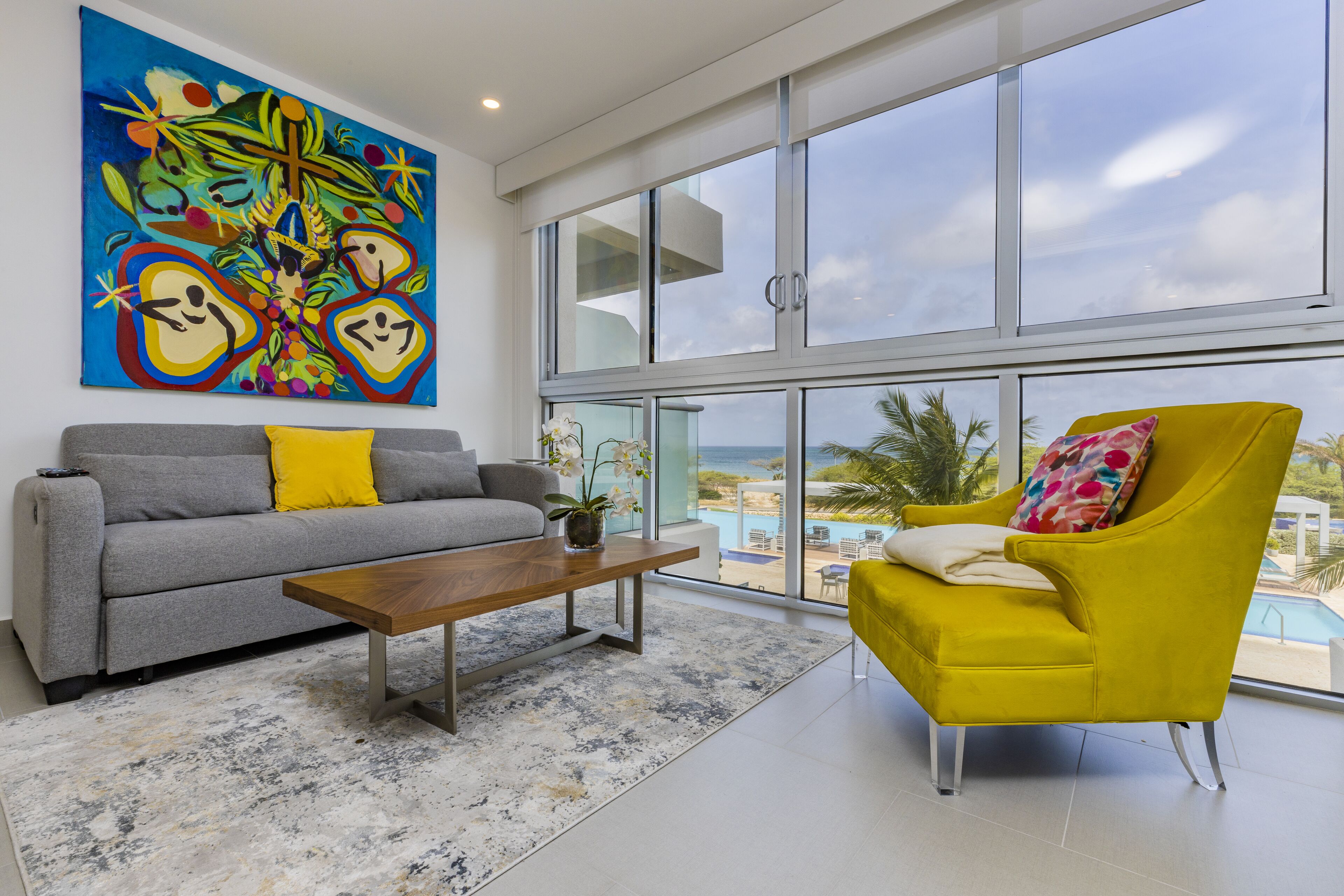 A modern living room with a gray sofa, a wooden coffee table, and a yellow armchair. A large abstract painting is on the wall, and there is a view of the ocean through large windows.