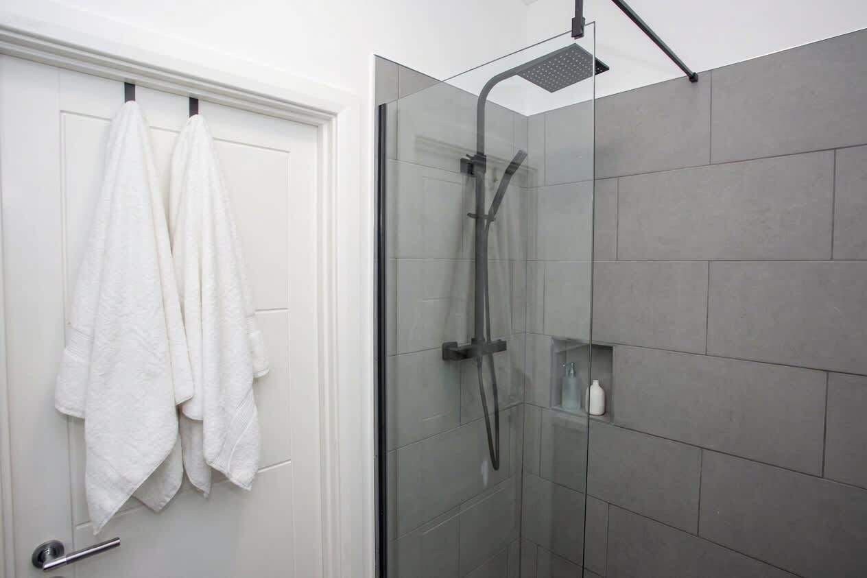 A bathroom with a glass-enclosed shower, white towels hanging on a door, and a small shelf with toiletries.