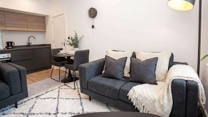 A modern living room with a sofa, a coffee table, and a lamp. There is a kitchen area with cabinets and a sink.