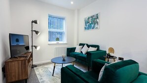 Een moderne woonkamer met een groene zithoek, een houten tv-meubel, een vloerlamp, een klein rond bijzettafeltje en een raam met zonwering.