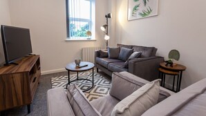 Uma sala de estar com sofá, mesa de centro, televisão em suporte de madeira, janela com persianas e uma planta sobre uma mesinha lateral.