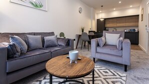 Uma sala de estar moderna com um sofá cinzento, uma poltrona cinzenta e uma mesa de centro redonda de madeira. Cozinha visível ao fundo, com armários de madeira e um micro-ondas.