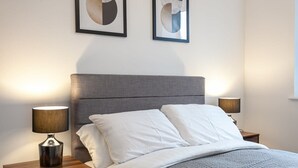 Cama bem arrumada com lençóis brancos, duas luminárias de cabeceira e arte abstrata na parede.