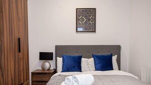 Un lit bien fait, avec des oreillers blancs et bleus, une table de chevet avec une lampe, et une œuvre d’art abstraite encadrée accrochée au mur.