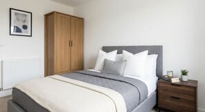 A bedroom with a bed, a wardrobe, a bedside table with a plant, and a framed picture on the wall. - Spacious & Modern 1 Bedroom Apartment in Bolton (Bolton)