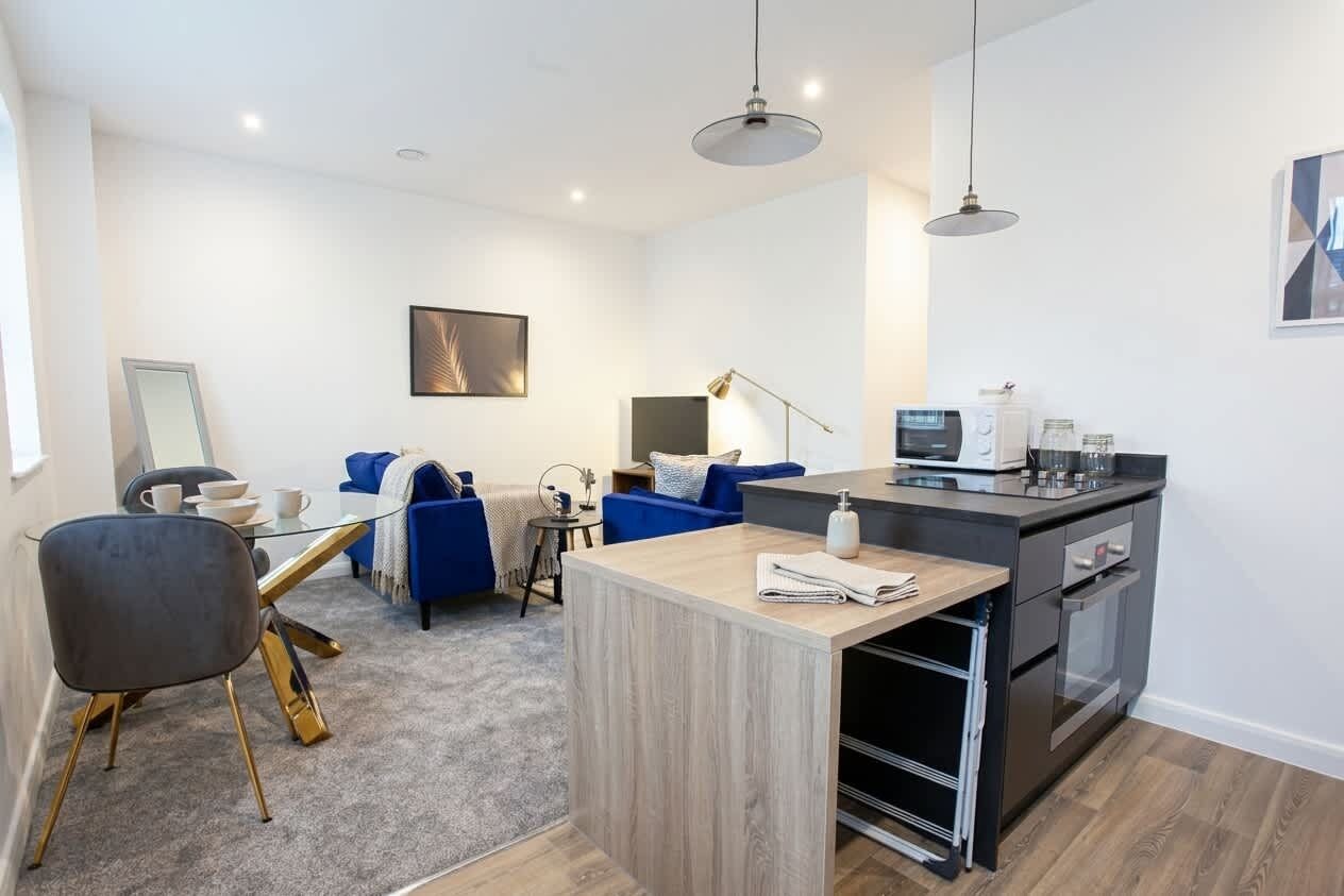 A modern kitchen with a built-in oven, a dining area with a glass table, and a living space with a sofa and a lamp.