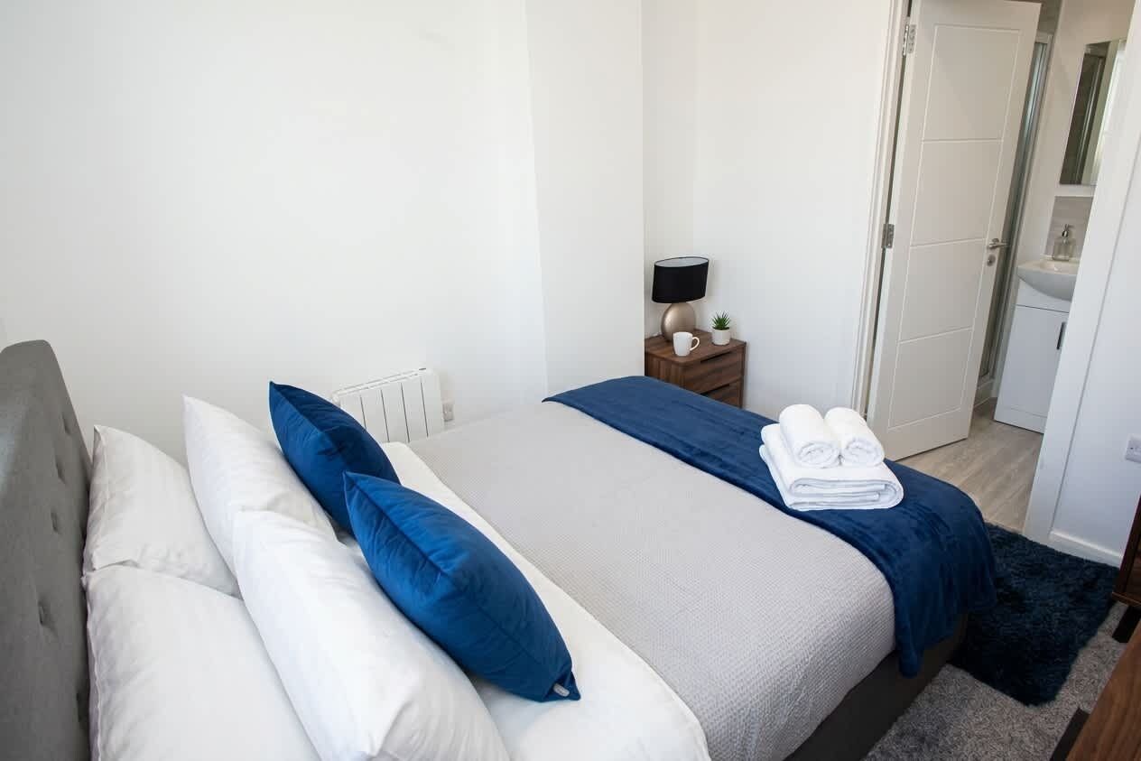 A neatly made bed with white and blue bedding, a bedside table with a lamp, and a door leading to a bathroom.
