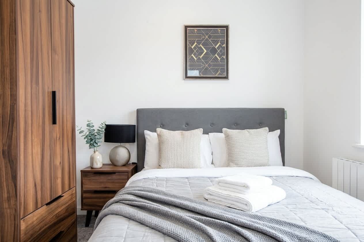 A bedroom with a bed, a nightstand, a lamp, a framed picture, and a wooden wardrobe.