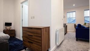 Une chambre avec une commode en bois, un lit, une table de chevet avec une lampe, un radiateur et une fenêtre donnant sur un immeuble.