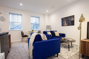 A modern living room with a blue sofa, a coffee table, and a TV. There are windows with blinds, a clock, and a lamp. - Spacious & Modern 1 Bedroom Apartment in Bolton (Bolton)