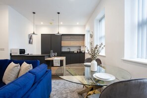 A modern living space with a glass dining table, a blue sofa, a kitchen area, and a window with curtains.