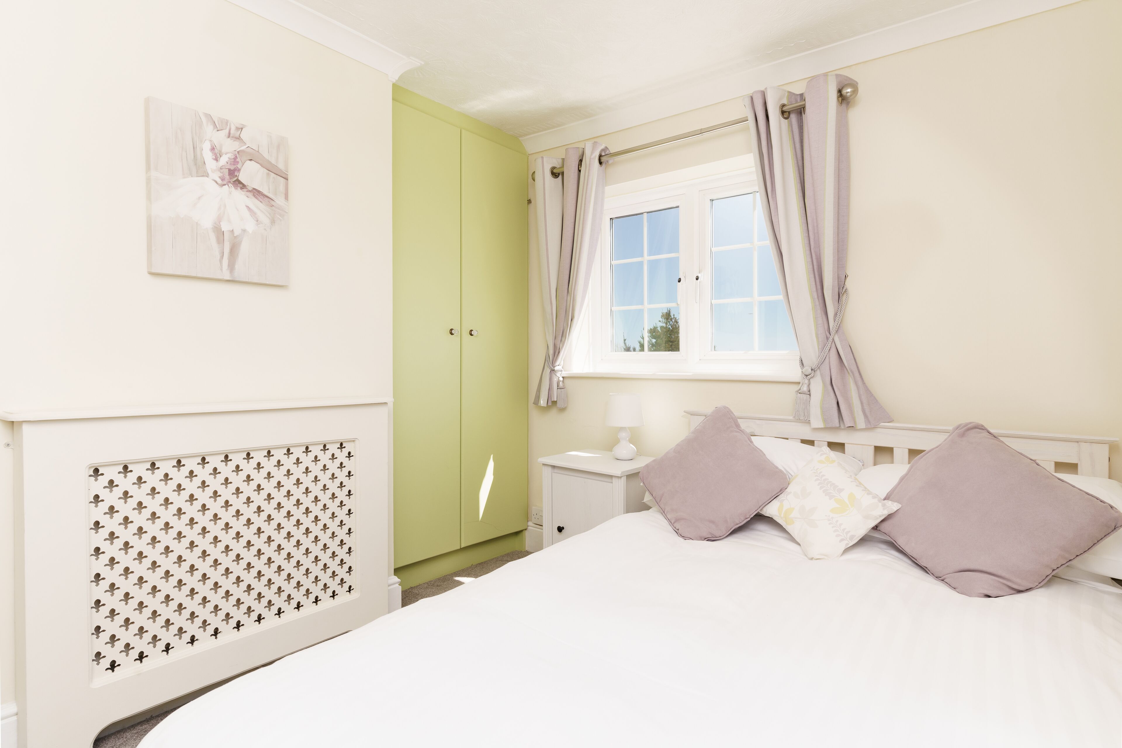 A bedroom with a bed, a nightstand, a window with curtains, a green wardrobe, and a wall-mounted radiator.