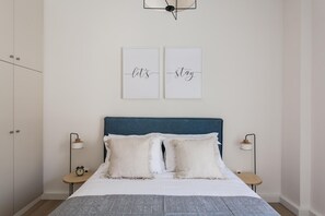 A bedroom with a bed, two bedside tables, a wall-mounted light fixture, and two framed pictures.
