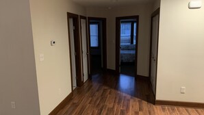 Un pasillo con suelo de madera, paredes blancas y una puerta que da a un dormitorio con cama y persianas.