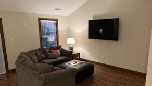 Una sala de estar con un sofá modular, un televisor de pantalla plana montado en la pared, una lámpara sobre una mesita auxiliar y una ventana con persianas.