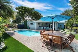 A poolside area with a dining table and chairs, a blue umbrella, and a house with a mural.