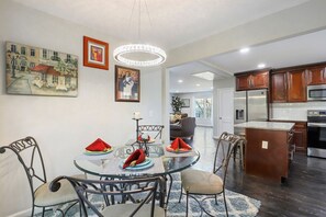 A dining area with a round glass table, metal chairs, and a chandelier. There are paintings on the walls and a kitchen in the background.