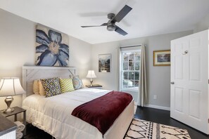 A bedroom with a bed, a ceiling fan, a window with curtains, and artwork on the walls.