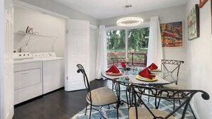 Una cocina moderna con electrodomésticos blancos, un comedor con mesa de vidrio y sillas metálicas, y vistas a un jardín a través de un amplio ventanal.