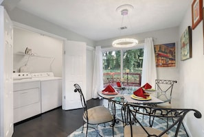 Cozinha moderna com eletrodomésticos brancos, área de jantar com mesa de vidro e cadeiras de metal, e vista para um jardim por uma grande janela.