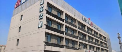 A multi-story hotel building with a prominent sign on the facade, multiple balconies, and a parking area with several cars.