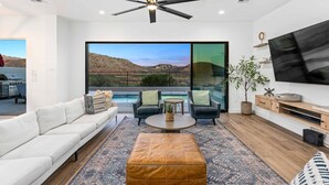 A modern living room with a white sofa, two armchairs, a coffee table, and a large TV. There is a ceiling fan, a large window with a view of a desert landscape, and a potted plant. - Double Eagle On The 8th: Private Pool + Near Coral Canyon (Washington)