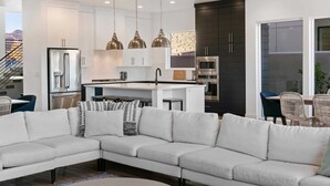 A modern kitchen with white cabinets, a stainless steel refrigerator, and a central island. There is a dining area with wicker chairs and a white sofa. - Double Eagle On The 8th: Private Pool + Near Coral Canyon (Washington)