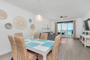 A modern living room with a dining area, featuring a white table with blue placemats, woven chairs, and a ceiling fan.